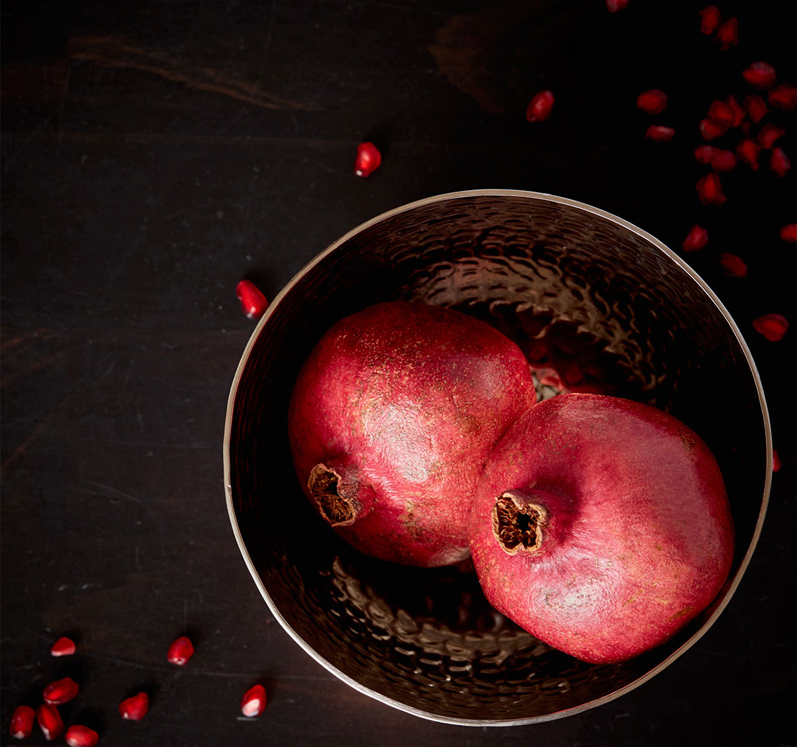 Pomegranates in bowl surrounded by pomegranate seeds photographed by Kate Benson, professional food and beverage photographer.